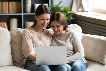 happy adult daughter and mature mother using laptop together, sitting on couch at home, smiling youn