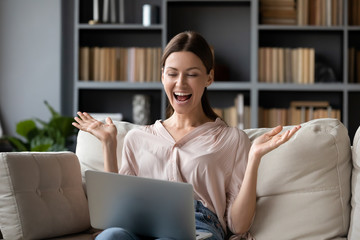 Excited woman looking at laptop screen, screaming with joy, surprised by message with good news, overjoyed young female sitting on couch at home, using computer, celebrating online win or success