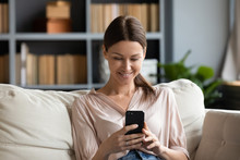 Smiling Young Woman Using Phone, Sitting On Couch At Home, Looking At Smartphone Screen, Beautiful Female Chatting In Social Network Or Shopping Online, Spending Leisure Time With Mobile Device