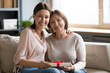 © fizkes - Portrait daughter hugging and congratulating older mother with birthday, mothers day or 8 march, happy young woman and mature mom holding gift box, looking at camera, sitting on couch at home