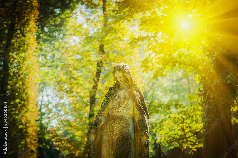 Ancient stone statue of Virgin Mary against beautiful landscape of forest in sunlight. - obrazy, fototapety, plakaty 