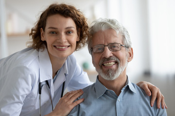 Wall Mural - Happy young adult female doctor physician wearing white coat and stethoscope embracing smiling healthy senior adult male patient looking at camera. Elderly people medical health care concept. Portrait