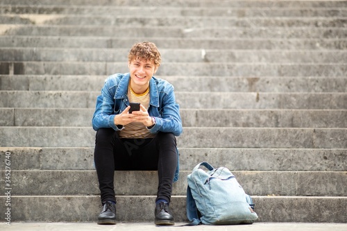Cuadros en Lienzo Full length smiling young man sitting on steps outside holding cellphone