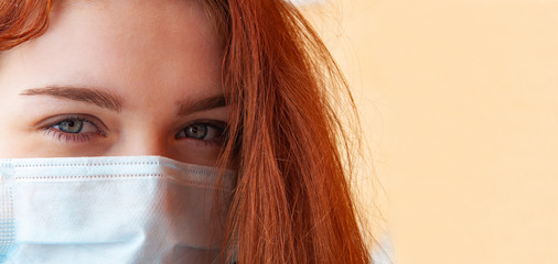 Close up horizontal shot of ill redhead grey eyes girl wears medical mask to protect from catching disease, stands outdoor, suffers from epidemic illness, yellow-brown background, copy space on right