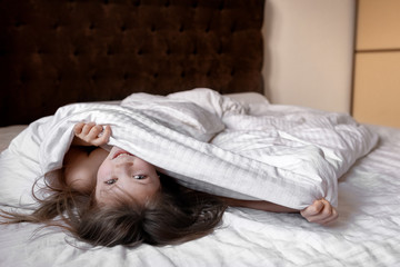 a little girl looks out from under the covers on the bed at home. portrait of a cute smiling little 