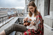 Young caucasian hipster/hippie woman in casual clothes playing on Hawaiian guitar, sings a song on a ukulele during self-isolation in the house on terrace. Hobby. Daily life in your own home. 