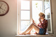 © pikselstock - Young woman sitting on window sill and using smartphone
