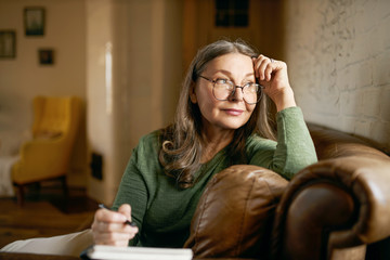 Stylish inspired middle aged woman writer wearing glasses sitting in leather couch with pen making notes. Attractive mature female artist sketching or teacher preparing tasks for online lessons