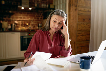 Indoor image of frustrated female pensioner managing finances at home, sitting at desk with portable computer, papers, paying bills online, having stressed look, trying to make both ends meet