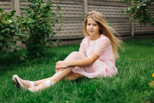 Beautiful Blue-eyed Girl With Long Blond Hair. Little Girl In A Pink Flamingo Dress. Summer Bright, Emotional Photo.  Girl Model With Long Hair Sits On Green Grass