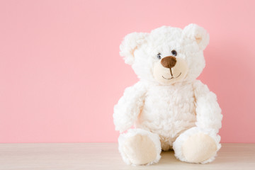 smiling white teddy bear sitting on table at pastel pink wall background. kids best friend. front vi
