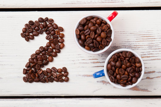 Top view two cups filled with coffee beans. Number two in a shape of heart. White wood on background.