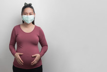 Asian pregnant woman wearing surgical mask looking at camera and holding her stomach by two hands, studio lighting portrait and copy space on grey background, covid19 concept
