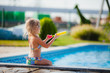 © Alina - Little blonde girl in a swimsuit sitting with her back to the camera and splashing water in the pool inn the hot summer