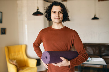 Positive young man in casual clothes holding roll of yoga mat under his arm going to practice indoors, happy to stay home during quarantine, spending more time on self development and healthy activity