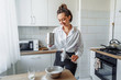 © Daria - DIY home cooking concept. A bright brunette woman sifting the flour for pancakes through a sieve, salting the dough, adding milk. Young woman smiling, loves to cook in a beautiful modern kitchen.