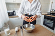 © Daria - A young beautiful woman in white clothes is preparing to cook in the kitchen, breaking egg in bowl. Cozy white kitchen with all equipment, prepared for cooking pancakes.