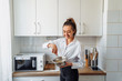© Daria - Beautiful, attractive, smiling young woman kneading dough with whisk for banana pancakes in steel dishes in a kitchen with white, light interior. Monochrome photo without bright accents