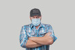 Caucasian man dressed casual with plain blue shirt and black baseball cap wears face medical hat and looks angrily at the camera. Mean looking man with crossed arms and safety mask isolated on gray. 