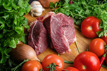 The preparation of fresh meat. Fresh beef steak on a wooden table. Meat surrounded by spices and vegetables. Fresh fruit. Red ripe tomatoes. Dietary food.