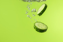 Falling Of Fresh Cucumber Slices Into Water Against Color Background