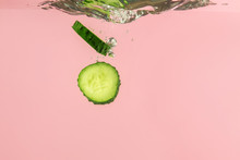 Falling Of Fresh Cucumber Slices Into Water Against Color Background