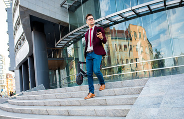 Wall Mural - Businessman with electric scooter walking in front of modern business building looking at phone.