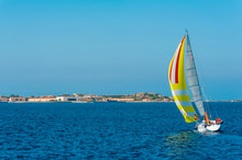 A View Of A Colorful Lonely Sailboat As It Sails Tilted By The Wind In The Mediterranean Sea With The Coast In The Background On A Sunny Day With The Blue Sky On Summer, In Sardinia Italy
