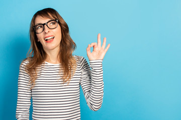 Portrait of a young friendly woman with a smile in a casual t-shirt and glasses makes an ok gesture and looks up on an isolated blue background. Emotional face. Gesture is all right, it's OK