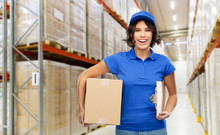 Logistics, Mail Service And Shipment Concept - Happy Smiling Delivery Girl With Parcel Box And Clipboard In Blue Uniform Over Warehouse Background