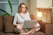 © Roman Tyukin - A young woman in a dress sitting on the couch with legs crossed works remotely on a laptop in her apartment. A girl during a webinar at home. A female student in glasses listening to an online lecture