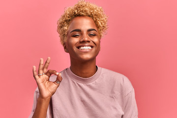 Gesture everything ok. Smiling black woman has all under control, gives positive reply, recommends good product, assure things are going well, likes something, looks gladfully at camera, stands indoor