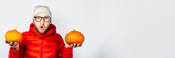 surprised young man in a red jacket and hat, holds two pumpkins on a light background. Concept of Halloween, celebration, autumn. Banner