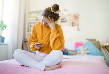 Wall Mural - Sad young female student sitting on bed, using smartphone.