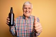 Senior handsome hoary man drinking bottle of beer standing over isolated yellow background happy with big smile doing ok sign, thumb up with fingers, excellent sign