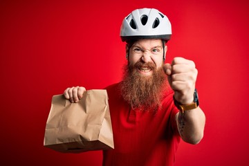 Redhead Irish delivery man with beard wearing bike helmet and holding takeaway paper bag annoyed and frustrated shouting with anger, crazy and yelling with raised hand, anger concept