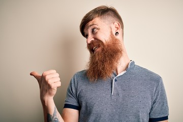 Sticker - Handsome Irish redhead man with beard and arm tattoo standing over isolated background smiling with happy face looking and pointing to the side with thumb up.