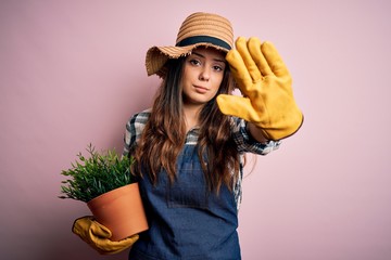 Wall Mural - Young beautiful brunette farmer woman wearing apron and hat holding pot with plants with open hand doing stop sign with serious and confident expression, defense gesture