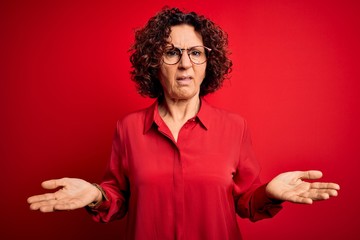 Poster - Middle age beautiful curly hair woman wearing casual shirt and glasses over red background clueless and confused with open arms, no idea concept.