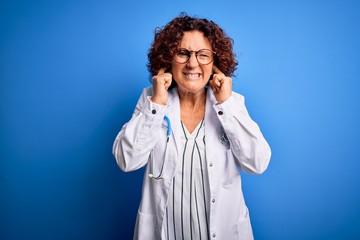 Sticker - Middle age curly hair doctor woman wearing coat and stethoscope over blue background covering ears with fingers with annoyed expression for the noise of loud music. Deaf concept.