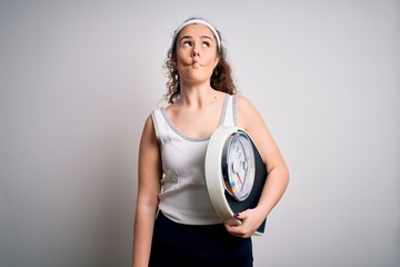 Wall Mural - Young beautiful woman with curly hair holding weighing machine over white background making fish face with lips, crazy and comical gesture. Funny expression.