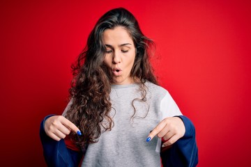 Wall Mural - Young beautiful woman with curly hair wearing casual sweatshirt over isolated red background Pointing down with fingers showing advertisement, surprised face and open mouth