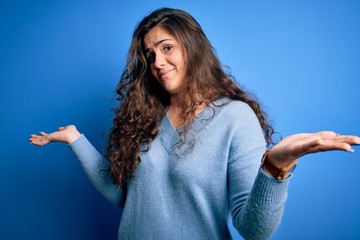 Sticker - Young beautiful woman with curly hair wearing blue casual sweater over isolated background clueless and confused expression with arms and hands raised. Doubt concept.