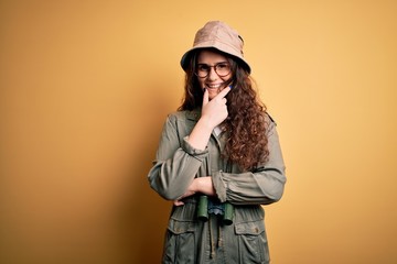 Poster - Young beautiful tourist woman on vacation wearing explorer hat and binoculars looking confident at the camera smiling with crossed arms and hand raised on chin. Thinking positive.