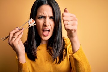 Wall Mural - Young brunette woman with blue eyes eating salmon maki sushi using chopsticks annoyed and frustrated shouting with anger, crazy and yelling with raised hand, anger concept