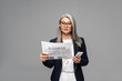 © LIGHTFIELD STUDIOS - attractive asian businesswoman in eyeglasses listening music with earphones and smartphone while reading business newspaper isolated on grey