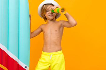 attractive European blond boy in a summer panama and yellow swimming trunks with an inflatable swimming board on an orange background