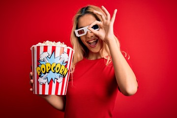 Sticker - Young beautiful blonde woman watching movie using 3d glasses eating popcorns as snack with happy face smiling doing ok sign with hand on eye looking through fingers