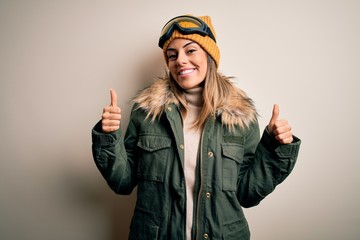 Poster - Young brunette skier woman wearing snow clothes and ski goggles over white background success sign doing positive gesture with hand, thumbs up smiling and happy. Cheerful expression and winner gesture