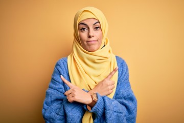 Sticker - Young beautiful brunette muslim woman wearing arab hijab over isolated yellow background Pointing to both sides with fingers, different direction disagree